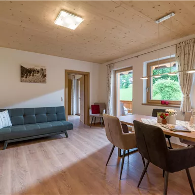 A modern living room with a sofa and a dining table. Large windows let in plenty of daylight and offer a view of greenery.