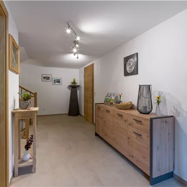 A modern hallway with bright walls and warm lighting. On the left side, there is a wooden chest of drawers and on the right side, a coat rack.