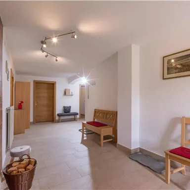 A bright hallway with wooden chairs and a bench. The walls are plain, and a picture hangs over one of the chairs.