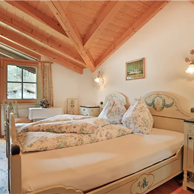 A cozy bedroom with slanted wooden beams and a large window. The room is furnished with a double bed and stylish furniture in a traditional design.