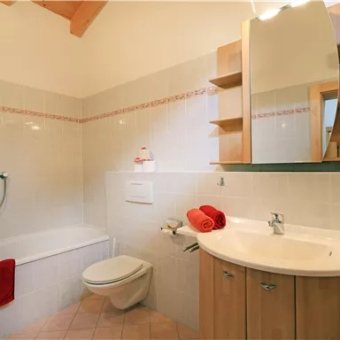 A modern bathroom with a bathtub and a shower. The room is bright and features wooden shelves as well as towels in warm shades.