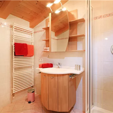 A modern bathroom with a wooden cabinet and a large mirror. The walls are light-colored tiles and there are towels in bright red.
