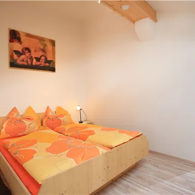 A cozy bedroom with a wooden bed and orange pillows. In the background, a bathroom is visible.