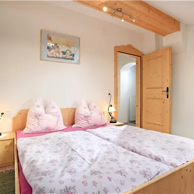 A cozy bedroom with a double bed and pink bedding. The walls are light, and there is a wooden door as well as a beautiful wall decoration.