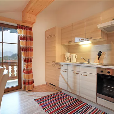A modern kitchen with light wooden cabinets and a large window. The view outside shows a picturesque mountain landscape.
