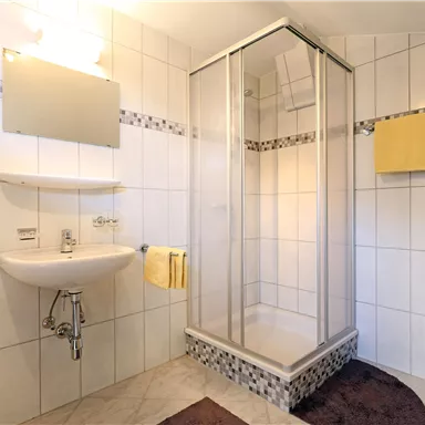 A modern bathroom with a shower and a sink. Yellow towels and a nearby mat complement the bright design.