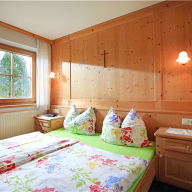 A cozy bedroom with wooden walls and a large window. The bed is decorated with colorful floral bedding and pillows.