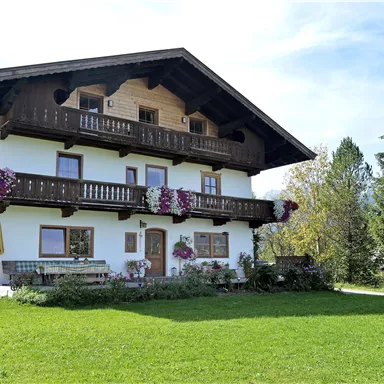 A charming two-story chalet with flowers on the balcony. In front of the house extends a green meadow and trees are visible in the background.