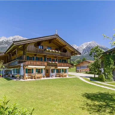 A traditional Alpine house surrounded by green grass and mountains in the background. The sky is clear and sunny.