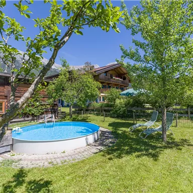 A beautiful garden with a round pool and a sunny lawn. In the background, you can see a wooden house and green trees.