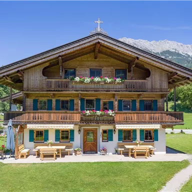 A traditional wooden house in alpine style, surrounded by green meadows and mountains in the background. Colorful flowers bloom on the balconies.