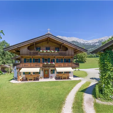 A traditional wooden house in a green landscape with mountains in the background. In front of the house, there are seating areas and a peaceful environment.