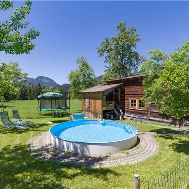 A beautiful garden with a round pool, green trees, and a cozy wooden house. In the background, mountain landscapes can be seen.