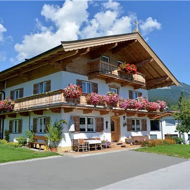 A charming house in alpine style with blooming balcony flowers. The surroundings are green, with mountains in the background and a blue sky.