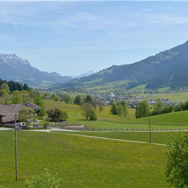 A picturesque landscape with green meadows and gentle hills. In the background, the mountains and a small village can be seen.