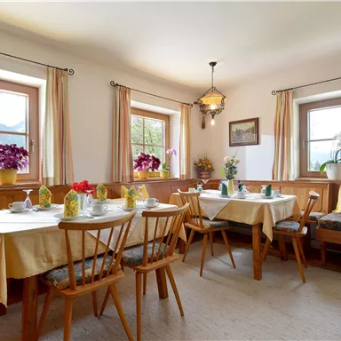 A cozy dining area with wooden furniture and tablecloths. Large windows offer a view of the mountains and fresh flowers decorate the room.