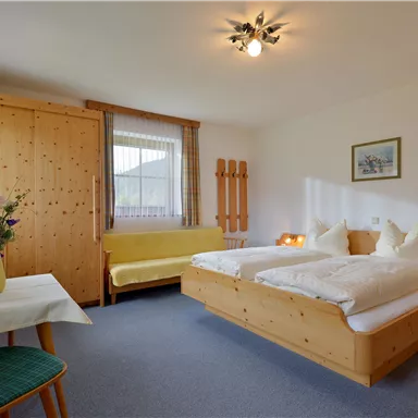 A cozy bedroom with two beds and a wooden wardrobe. There is a small table with a flower vase and a seating area.