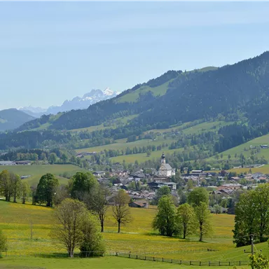 A picturesque landscape with green meadows and gentle hills. In the background, mountains and a small village are visible.