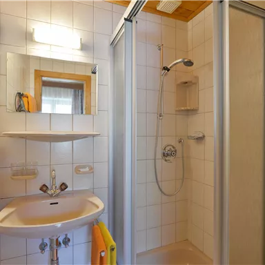 A small, bright bathroom with a shower, sink, and mirror. The walls are tiled, and there is a towel.