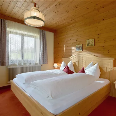 A cozy bedroom with a wooden bed and white pillows. The walls are made of wood and there are large windows that let in plenty of daylight.