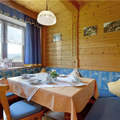 A cozy dining area in a wooden room with a table and blue seat cushions. The walls are made of wood, and the window lets in plenty of daylight.
