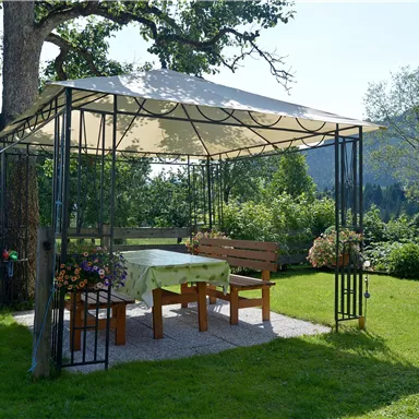 A shady pavilion with wooden furniture stands on a green meadow. In the background, trees and gentle hills can be seen.