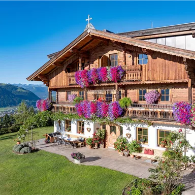 Ein traditionelles blaues Holzhaus mit bunten Blumen an den Balkonen. Im Hintergrund sind grüne Berge und ein klarer Himmel zu sehen.