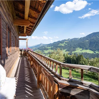 Ein Holzbalcon mit Blick auf grüne Hügel und Berge. Der Himmel ist klar und die Sonne scheint.