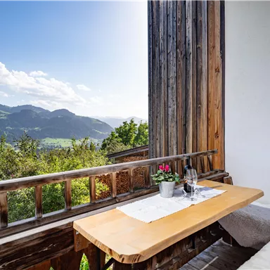 Ein gemütlicher Balkon mit einem Holztisch und Blick auf die Berge. Die Landschaft ist grün und lädt zum Entspannen ein.