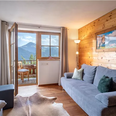 Gemütliches Wohnzimmer mit grauem Sofa und Blick auf die Berge. Große Fenster führen auf einen Balkon mit einem schönen Ausblick.