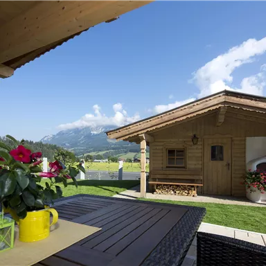 A cozy wooden house in a picturesque landscape. The table is decorated with flowers and colorful pots, and the sky is clear and blue.