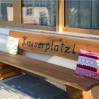A wooden bench with the inscription "Kaiserplatzl" stands in front of a window. On the bench, there are two decorative cushions.