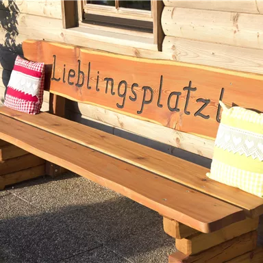 A wooden bench with the inscription "Favorite Spot" and colorful cushions. The background features a wooden facade and plants.