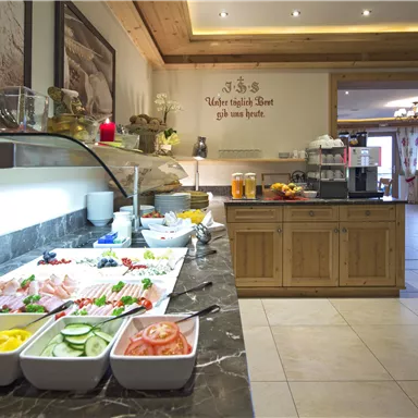 A rich breakfast buffet with a variety of fresh foods, including fruits, vegetables, and cold cuts. The room is bright and inviting with wooden ceilings and a cozy atmosphere.