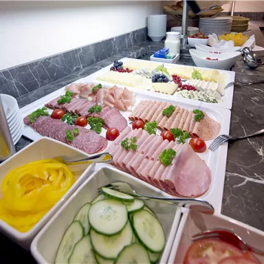 A hearty breakfast buffet with various types of cured meats and cheeses. Next to it, fresh vegetables and sauces on a standing table.