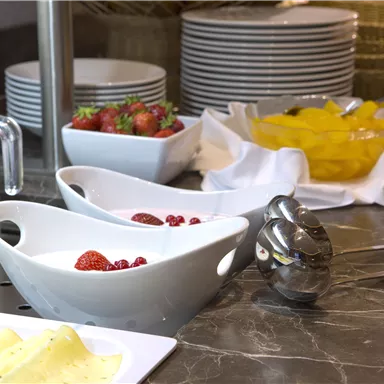 A breakfast table with fresh fruits, yogurt, and milk. In the background, additional dishware and fruit bowls are visible.