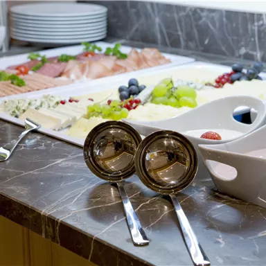 A beautifully arranged buffet table with various foods such as fish, fruit, and cheese. In the foreground, there are two shiny bowls and spoons visible.