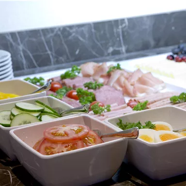 A buffet with fresh ingredients, including sausages, cheese, and various types of vegetables. Colorful bowls with cucumbers, tomatoes, and eggs complement the selection.