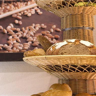 A basket with fresh bread and rolls is on a table. In the background, there is a painting of grains.