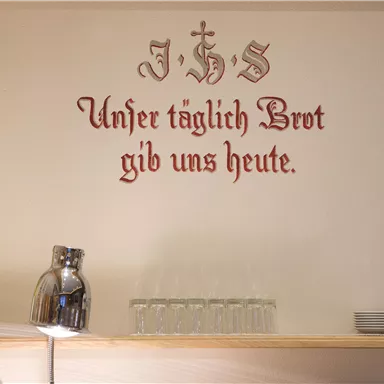 A wall with the inscription "Give us this day our daily bread" in decorative lettering. Next to it stands a jug and some cups.
