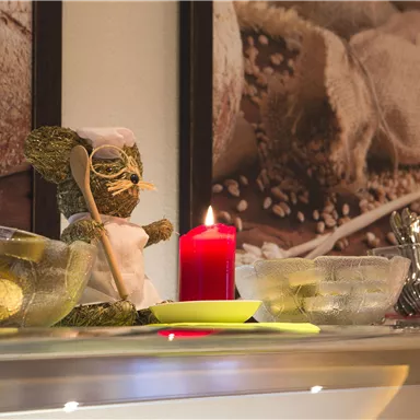A decorative kitchen counter with a small figure of a chef, a red candle, and several transparent bowls. In the background, there are pictures of bread and grains.