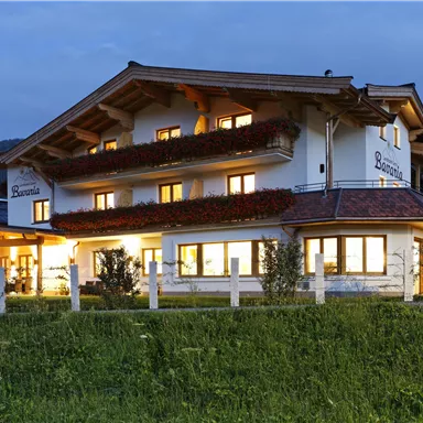 A cozy house with wooden cladding, illuminated at dusk. Surrounded by green meadows and mountains in the background.