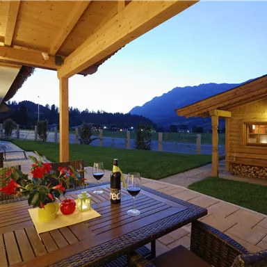 A cozy terrace with a wooden table and wine glasses. In the background, the mountains and a wooden house can be seen at dusk.