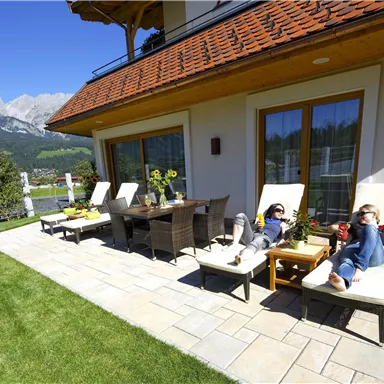 A quiet terrace with loungers and an outdoor dining table. In the background, green meadows and mountains are visible.