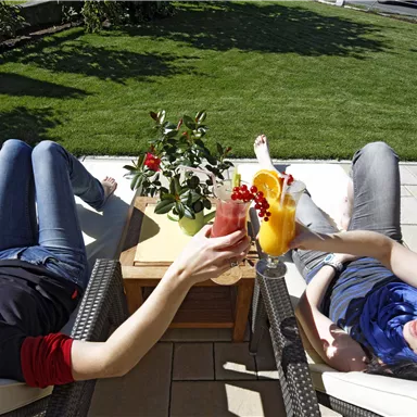 Two women relax on loungers outdoors and toast with colorful cocktails. The lawn is green and the atmosphere is sunny and relaxed.