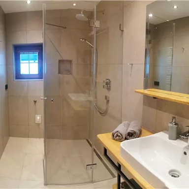 A modern bathroom with a glass shower and large tiles. There is a wooden shelf with various toiletries and towels.