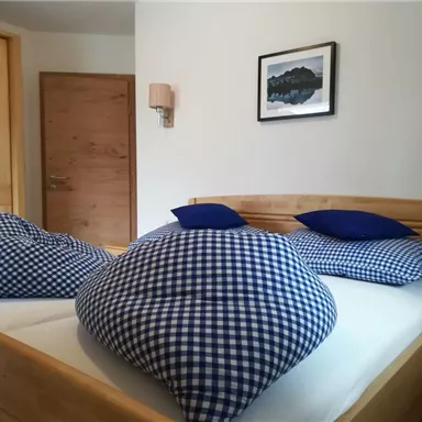 Ein gemütliches Schlafzimmer mit einem Doppelbett und großen karierten Kissen. Die Wände sind hell und es gibt einen Holzschrank sowie ein Bild an der Wand.
