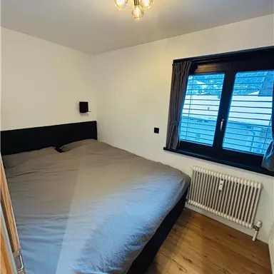 A simple bedroom with a large bed and a window. The walls are light and the floor is made of wood.