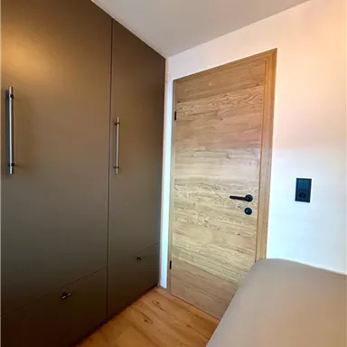 A modern bedroom with a wood wardrobe and a light wood door. The floor is wooden and the walls are kept neutral.