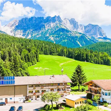 A picturesque landscape with a hotel building in the foreground. In the background, high mountains and green meadows can be seen.